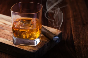 A glass of whiskey sits atop a wooden cutting board next to a lit cigar, which is producing wisps of smoke.
